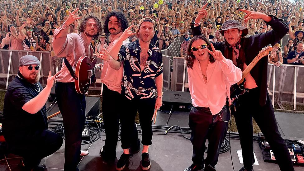 CVC band members. From left to right, Ben wearing glasses, a hat and navy t-shirt and a silver chain, crouching to the floor, Elliot with a brown guitar and pale shirt and black trousers with his right hand making a rock sign, Francesco wearing a pale shirt and black trousers with his left hand pointing to the camera, Tom with a black half shirt and white checked patters and black trousers, wearing a cap back to front making a rock sign with his right hand, Nanial wearing a pink shirt and dark trousers with sunglasss, with a peace sign from his left hand and Dave wearing a round hat, dark shirt and neck scarf, with his eyes closed and both hands up in the air, with hundreds of fans in the background.