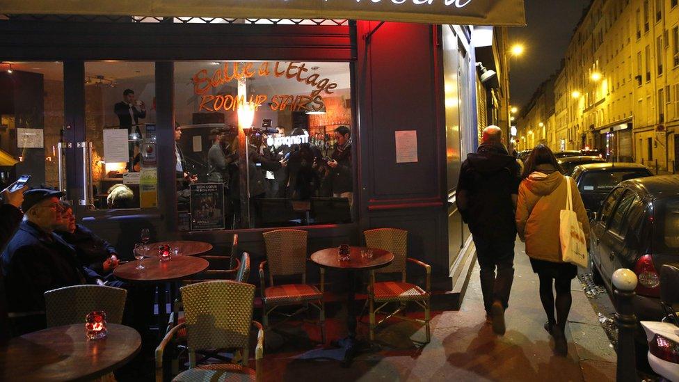 Two people walk down the path outside a restaurant, while two more sit on the exterior seating of the cafe / restaurant in Paris