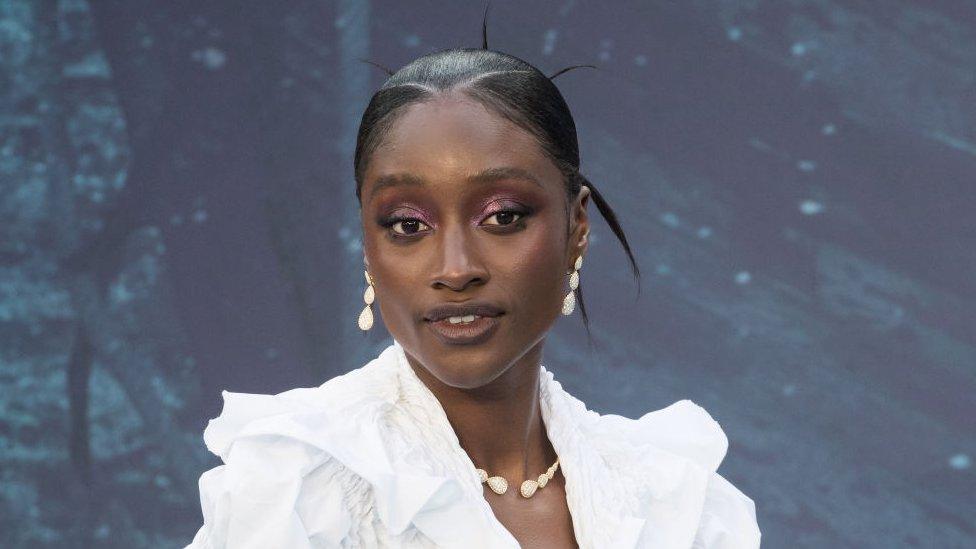 Sienna King on the red carpet, wearing a white blouse with open neck and puffy shoulders. Her hair is tightly tied back, and she wears dangly earrings made up of three teardrop-shaped stones. A matching necklace can also be seen.