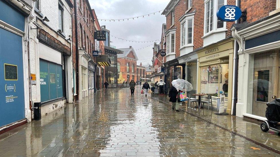 A view up a wet high street in Lincoln with shops on either side and people with umbrellas up