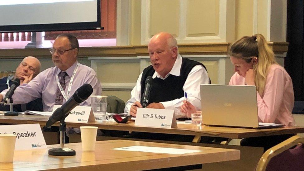 Councillors sitting at a desk. The middle one is wearing a black sleeveless sweater and white shirt.