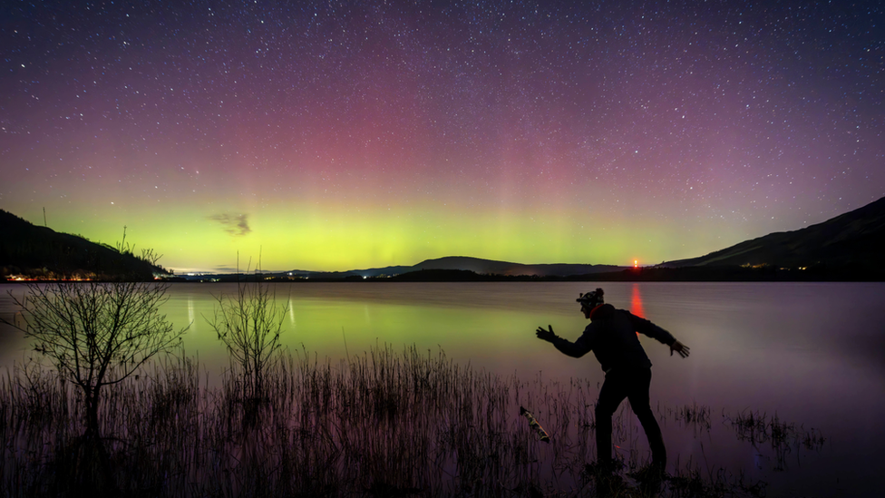 View across a lake with purple and green skies of the aurora in the background and a silhouette of a person in the foreground.