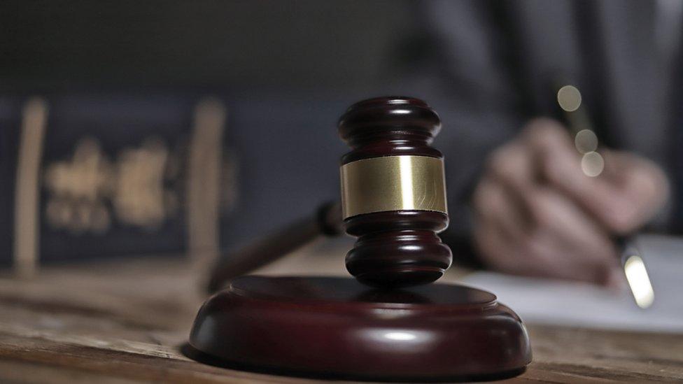 A gavel sitting on its round base, with a person visible behind, partially blurred out and writing on a desk