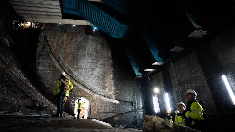 Engineers wearing yellow jackets and hard hats gaze at the underside of the bridge from within a chamber