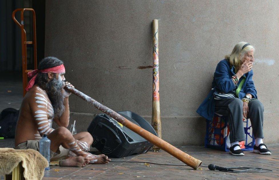 An Aboriginal man plays a didgeridoo while an elderly woman smokes a cigarette