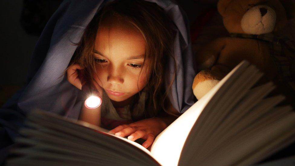 A young girl reads a book with a torch at night.