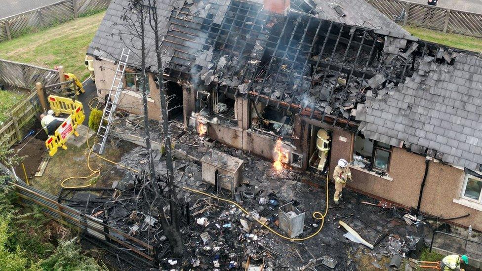 Image of firefighters at work on row of houses with burned roofs and yards