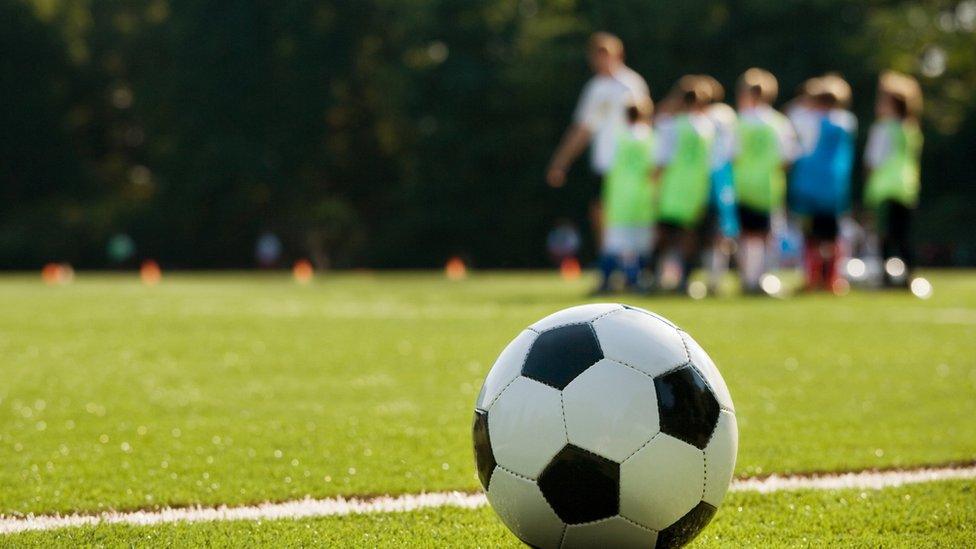 A football sits in the foreground on a white line while children take part in a coaching session in the background