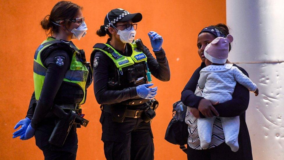 Two police officers speak to a woman carrying a baby out the front of a locked-down housing tower