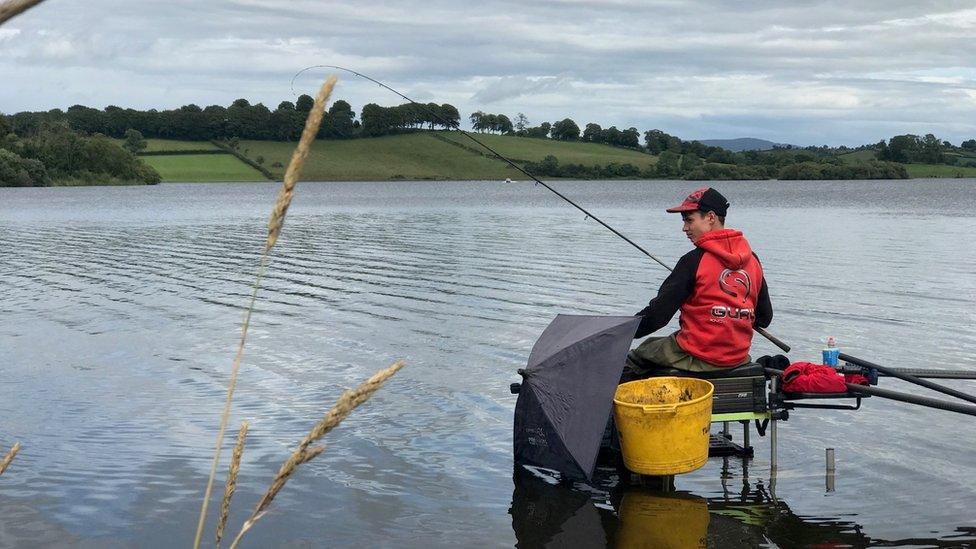 James fishing at the side of a lake, he is sitting in a chair and holding a rod