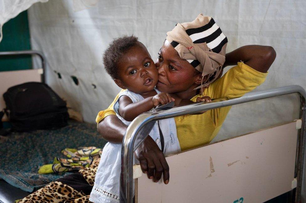 A infant sits in a hospital ward with her mother.