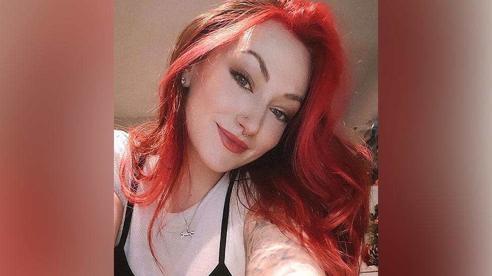 A woman with long, dyed red hair looks down into the camera. She's got red lipstick in a similar shade to her hair and black eyeliner. She also has a nose stud in her nostril and ring piercing in her septum. She's smiling, and wearing a white fitted t-shirt under a black strappy top. The top of her arm, which has an artistic black and white tattoo on it, is also visible.