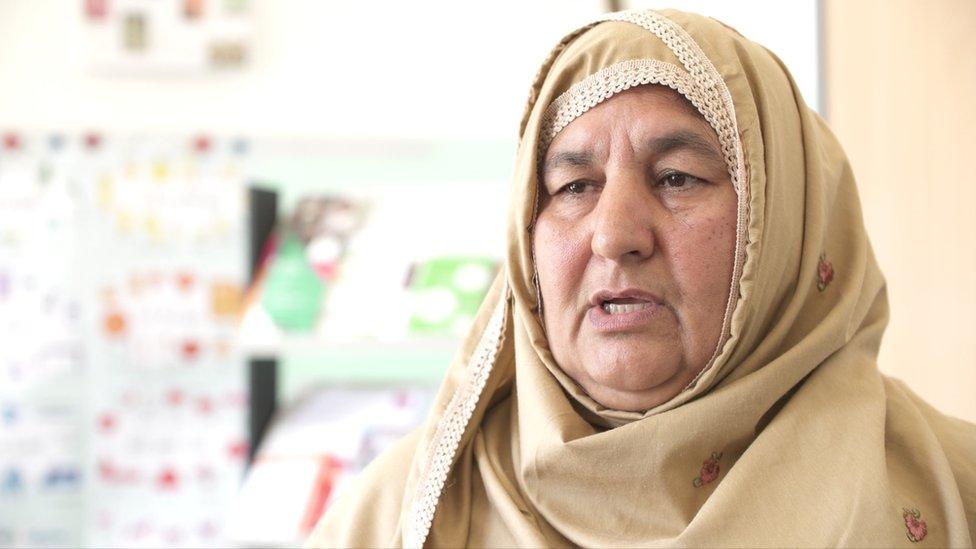 A woman is seen wearing a scarf around her head, with her face visible, as she speaks towards the camera