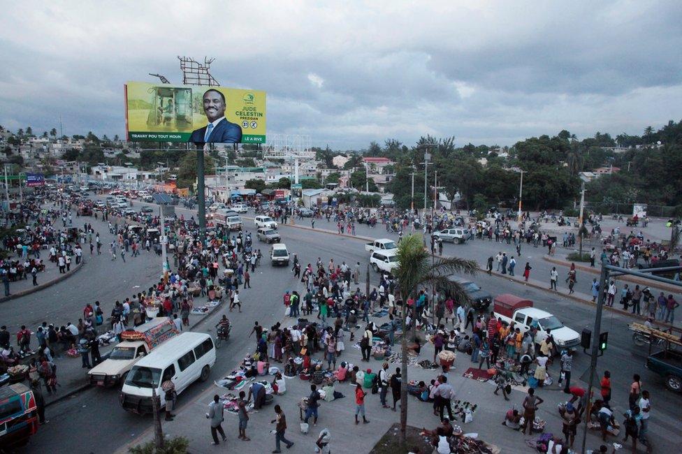 People walk in the street next to an electoral billboard of presidential candidate Jude Celestin in Port-au-Prince, Haiti, 17 November