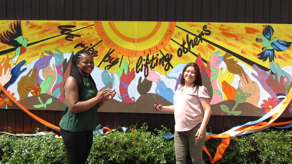 Sheila and Patricia, guests of The Harbour Project, smiling and clapping as they cut the ribbon lying across the mural