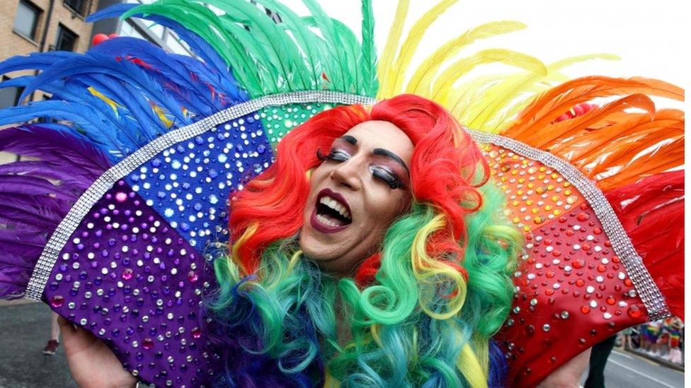 A marcher wears an oversized feather collar featuring the colours of the rainbow flag