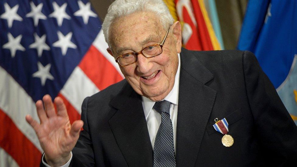 Henry Kissinger waves at the camera.