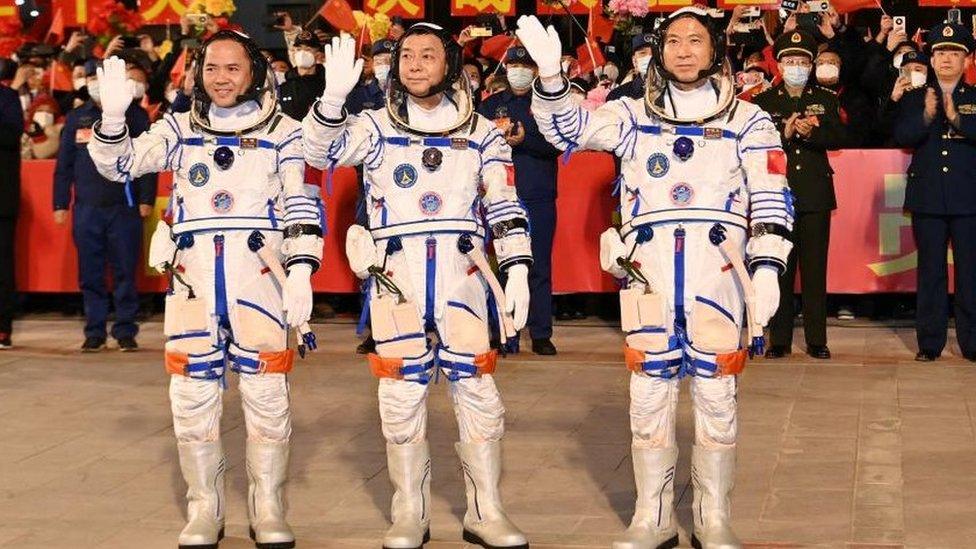 Three astronauts stand in their suits in front of Chinese officials and wave