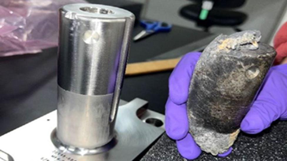 A scientist holds an eroded object that fell from space next to a shiny metal cylinder, which is how it looked when attached to the ISS.