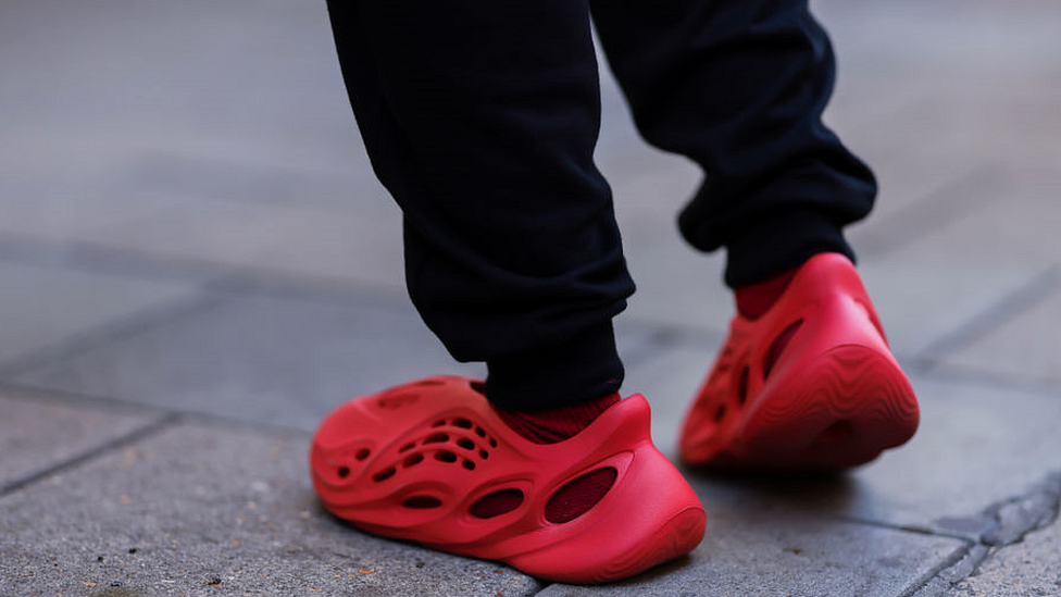 The bottom of a person's legs - they're wearing jogger-style trousers. They're wearing a pair of red Yeezy Sandals, which cover the whole foot but have large airholes dotted around the shoe. They contrast starkly against the grey paved surface they're walking on.