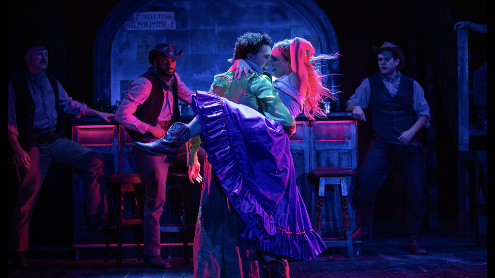 Brightly lit theatre stage. Two people dancing in colourful cowboy themed outfits