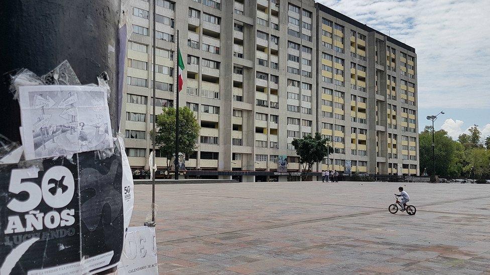 A boy cycles across the Plaza de las Tres Culturas in 2018, a poster about the student protests in the foreground says '50 years fighting'