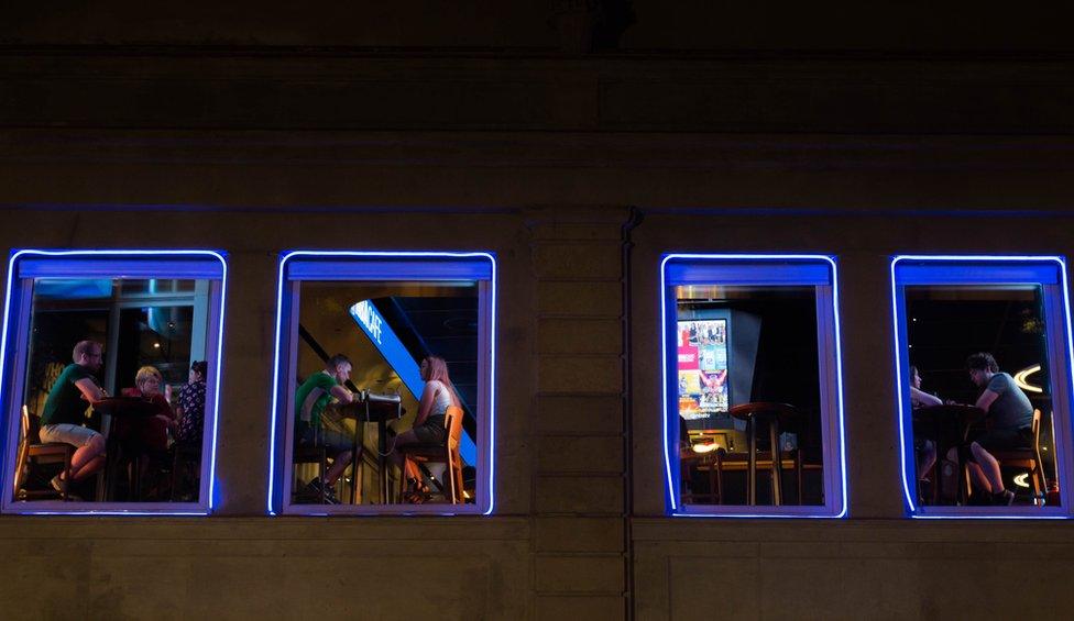 Through the windows of a bar, people are sat talking and drinking