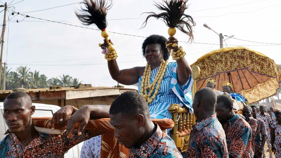 Queen is carried on a litter with leopard skin umbrella in the background giving shade to the king