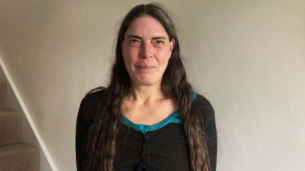 Image of Sue. She is stood against a cream wall and looking directly at the camera with a slight smile on her face. She has long dark hair and is wearing a dark t-shirt.