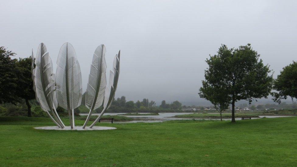 a stainless steel sculpture of huge eagle feathers in a circle, next to a tree on a cloudy grey day