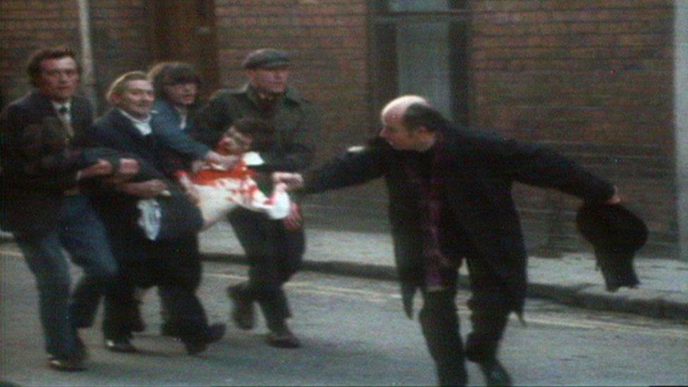 A priest waves a bloodstained handkerchief as four men carry an injured, bloodied man through the streets of Londonderry on Bloody Sunday