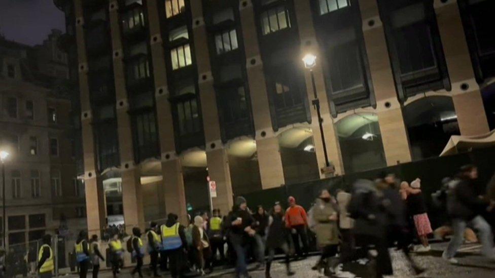 Dozens of people push past barriers manned by stewards wearing fluorescent vests.