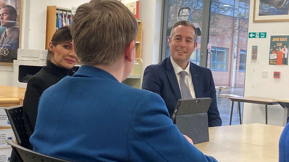 Paul Givan sits at a table speaking to a Rathmore pupil in a blue blazer