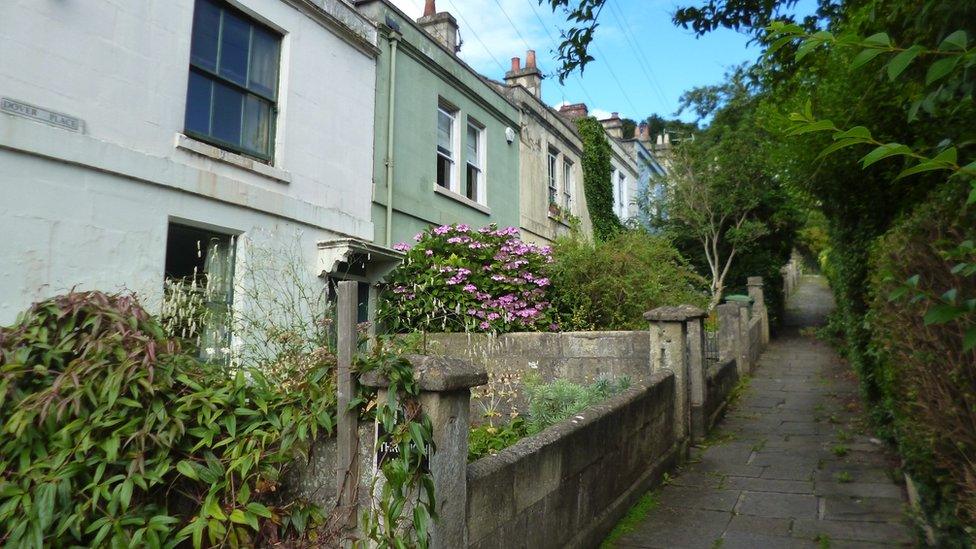 Photograph of Dover Place in Bath. It is a pedestrianized street lined with colourful cottages on the left, and trees on the right.