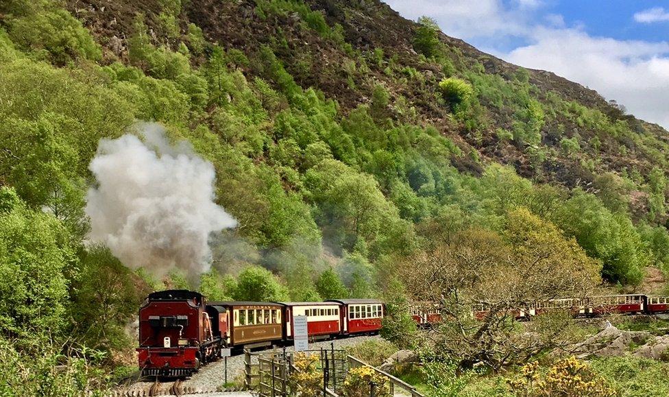 A photograph an old steam train on a railway. It's purple in colour and there's is a plume of white smoke coming from the funnel. In the background there is a rugged Welsh green hillside and a blue sky, with a couple of fluffy white clouds