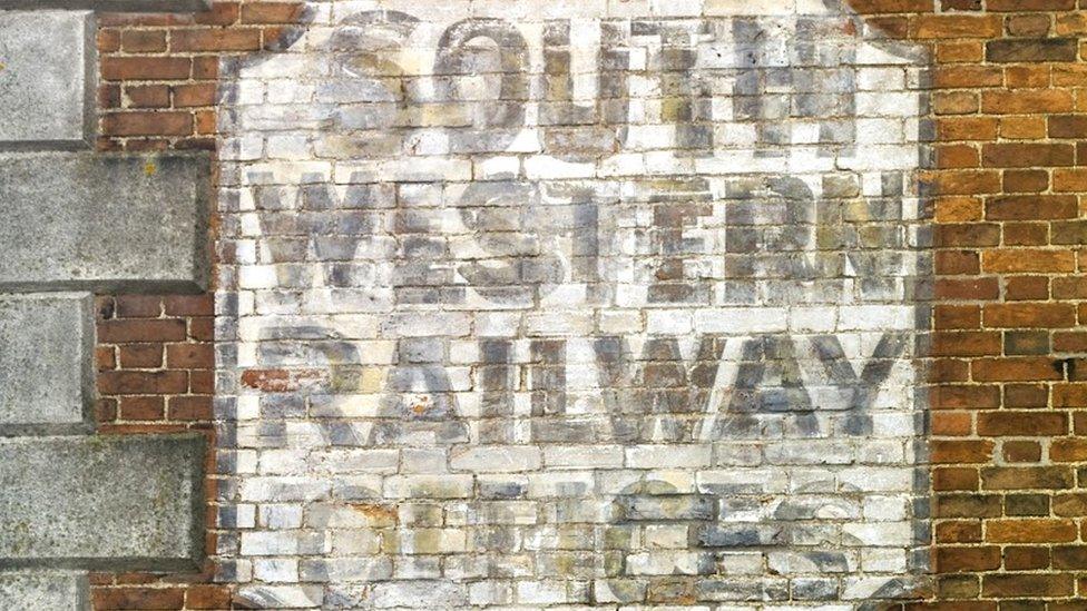 A faded painted sign on a red brick wall that has a while background and blakc lettering