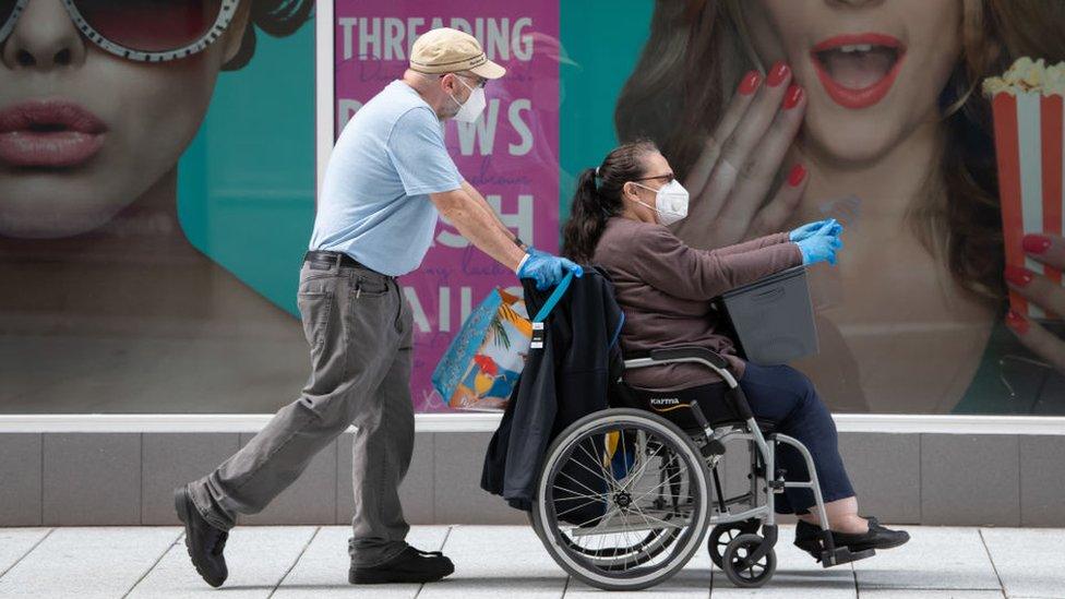 A woman sitting in a wheelchair wearing a mask and gloves, being pushed by a man wearing the same