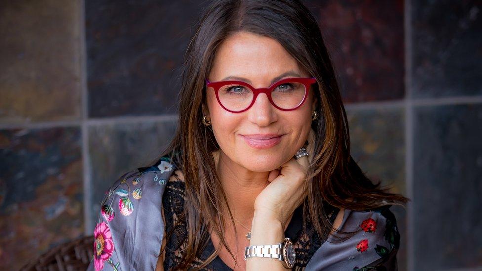 Ms Gersh, wearing red glasses and resting her head on one hand, looks at the camera in this portrait