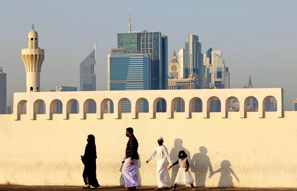 A family walk past a white wall with the skyline of Dubai in the background