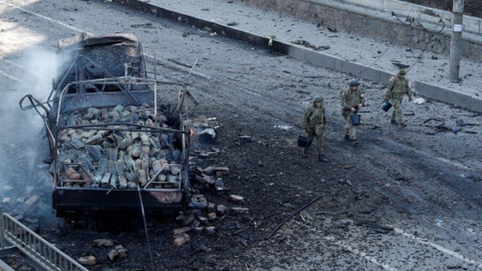 Ukrainian servicemen walk past a damaged vehicle in Kyiv