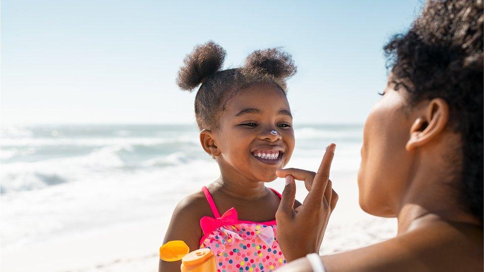 a girl at the beach in a swimming costume smiles as an adult put sunscreen on her face