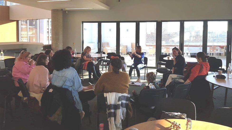 Image of a Mothers in Music session. The group of women are sat in a circle. A few of them can be seen smiling, and Alice can be seen talking, with her arms stretched out.