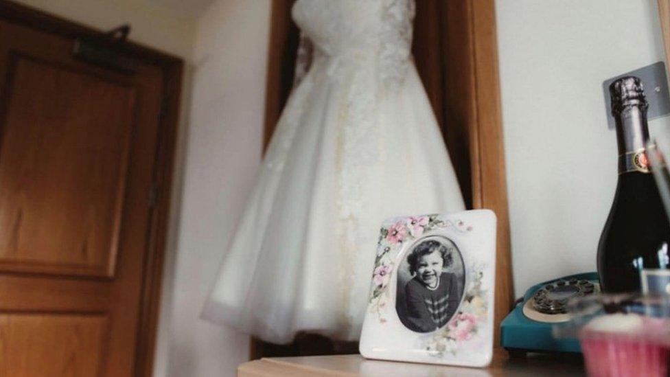 A photograph of Katrice on a dressing table with Natasha's wedding dress hanging in the background