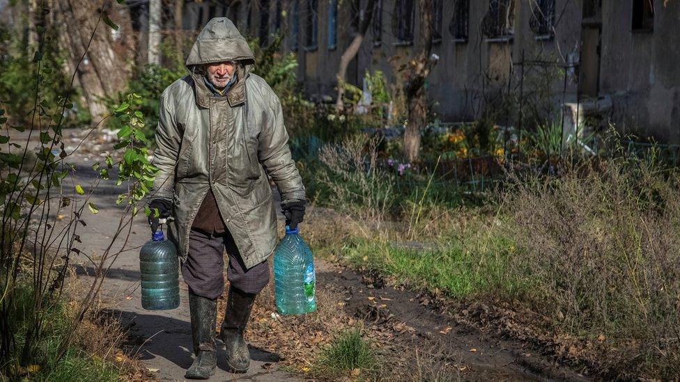 Man carrying two large bottles of water walks on a path in front of a damaged building