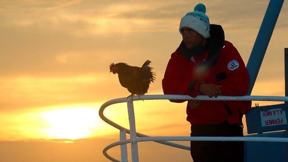 Sailor Guirec Soudee and hen Monique standing on the bow of a boat at sunset