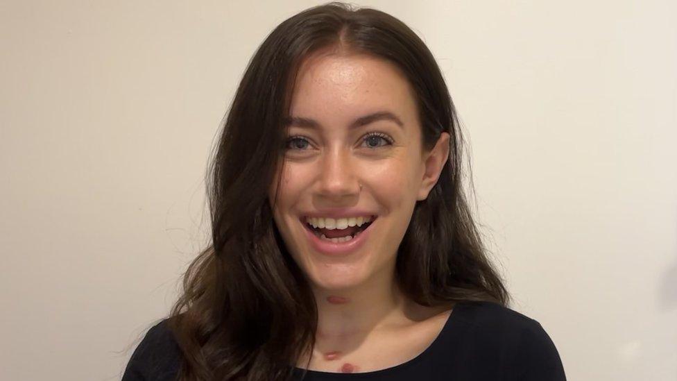 Natalie has straight brown hair and blue eyes and is smiling for the camera. She's wearing a black t-shirt and the scars from her cancer treatment are visible on the front of her neck