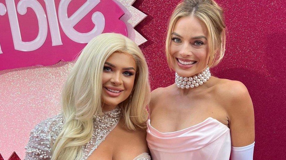 Liberty Poole and Margot Robbie, smiling at the camera. Liberty is wearing a silver dress and Margot is in a pink dress