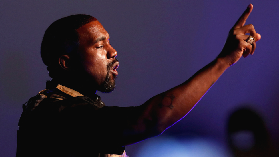 Kanye West appears to be addressing an audience. He has one arm raised up, his index finger pointing skywards, and looks determined as he speaks. There's a dark blue light emanating from the background, and a faint blue glow can be seen on the underside of his arm where his skin meets the light.