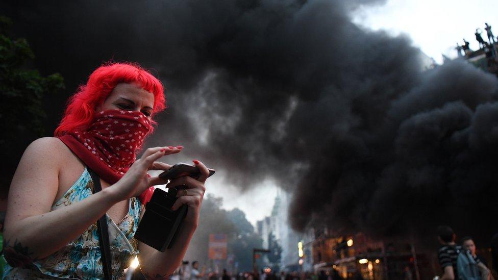 A demonstrator reviews a photo on her phone while a fire burns in the middle of town