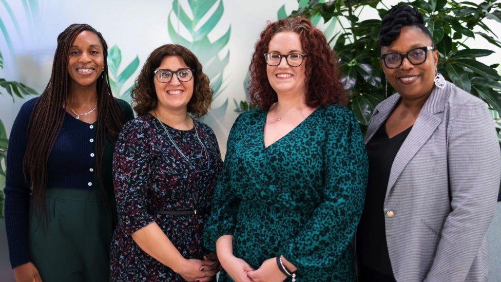 (L-R) Sonia Cohen, Kristal McNamara, Sarah Byfield and Sandra Gordon. They are stood in a line, smiling at the camera. There are plants and a white wall behind them.